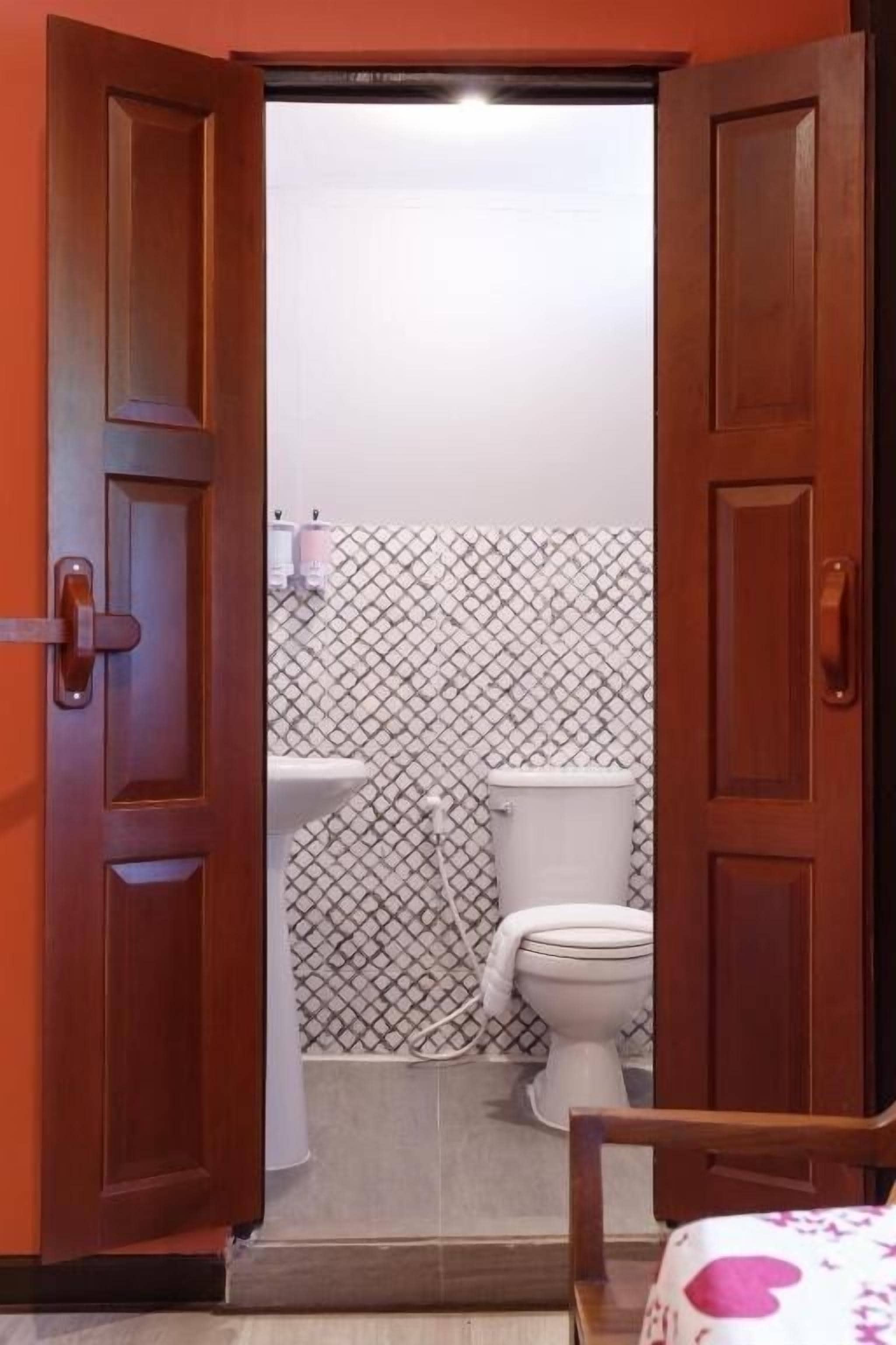 Eine geöffnete Holztür, die zu einem Badezimmer mit Toilette, Waschbecken und gemusterten Wandfliesen führt.