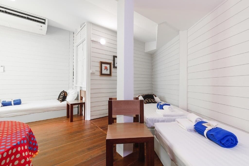 A small, cozy room with a wooden floor, a single bed with white bedding and blue towels, a wooden chair, a small table, and a bench with cushions.