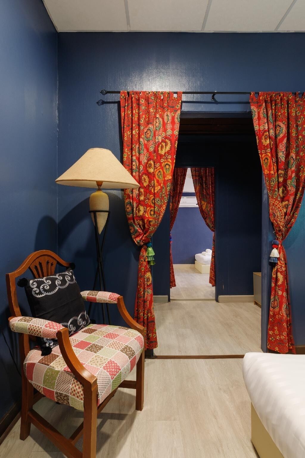 Une pièce comprenant une chaise en bois, une table, une lampe et un lit. Il y a des rideaux à motifs floraux.