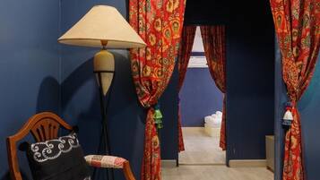 Une pièce comprenant une chaise en bois, une table, une lampe et un lit. Il y a des rideaux à motifs floraux.