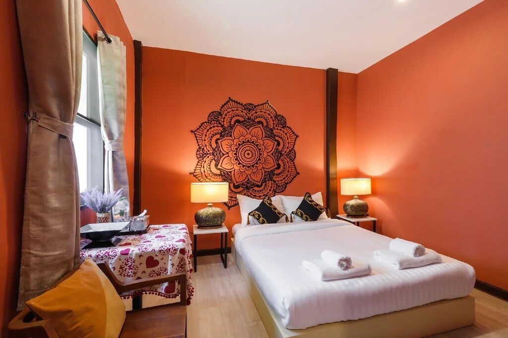A hotel room with a large mandala wall art, a bed with white linens, two bedside tables with lamps, a small table with a floral tablecloth, and a window with curtains.