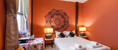 A hotel room with a large mandala wall art, a bed with white linens, two bedside tables with lamps, a small table with a floral tablecloth, and a window with curtains.