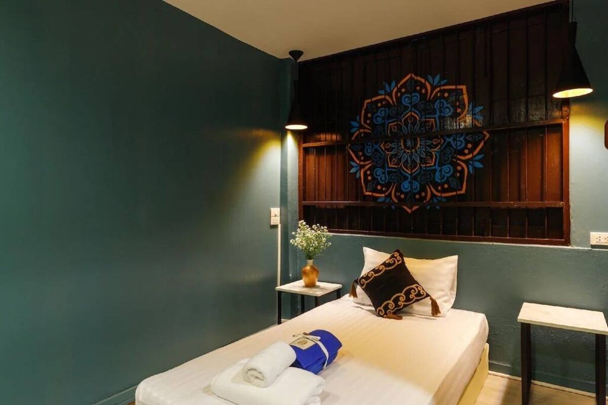 A hotel room with a bed, a wall with a decorative pattern, a small table with a vase, and a nightstand.