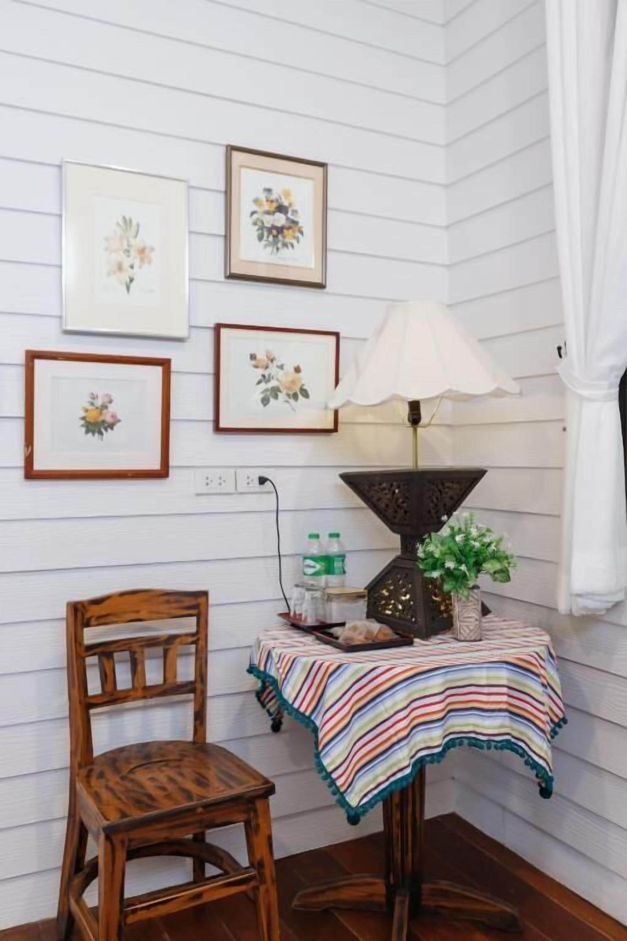 A corner room with white paneled walls, a wooden chair, a small table with a striped tablecloth, and framed floral artwork.