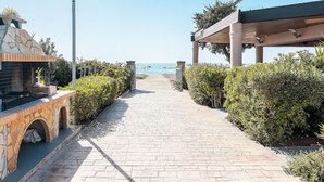 A paved pathway leading to a beach with a barbecue area and a covered seating area.
