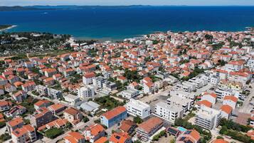 An aerial view of a coastal town with numerous buildings, some with red roofs, and a clear view of the sea and distant mountains.