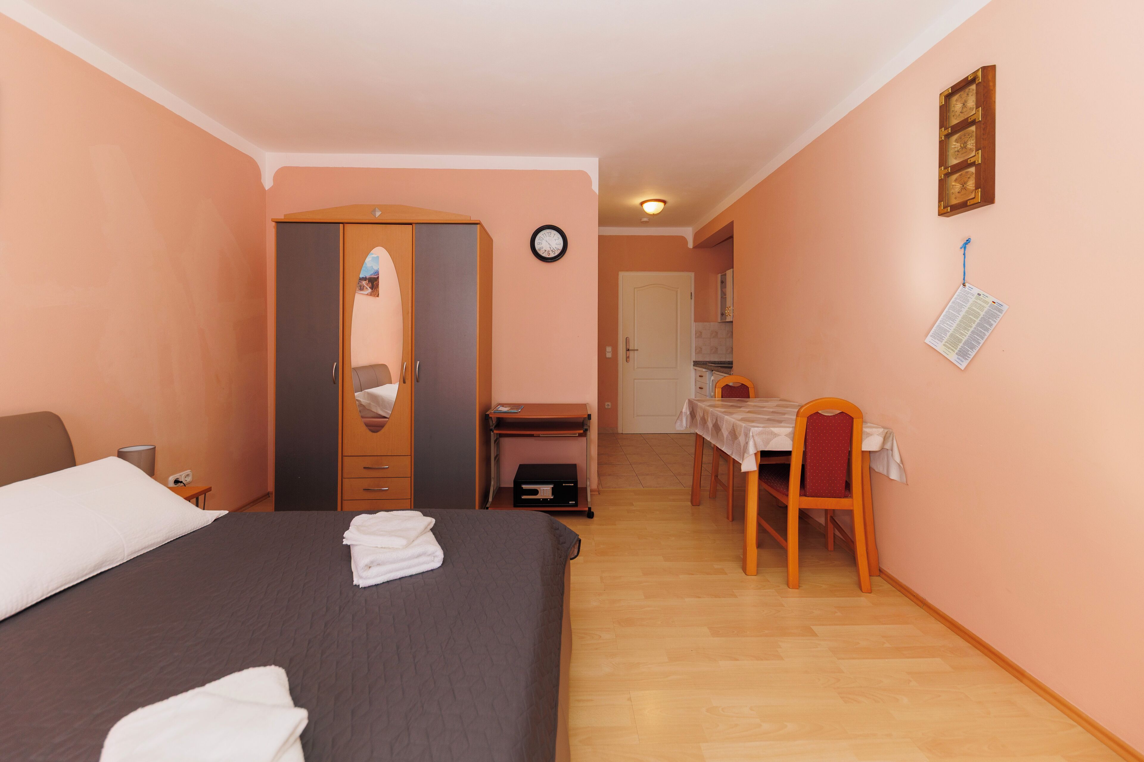 A hotel room with a bed, a desk, and a chair. There is a wardrobe, a clock, and a wall-mounted paper holder.