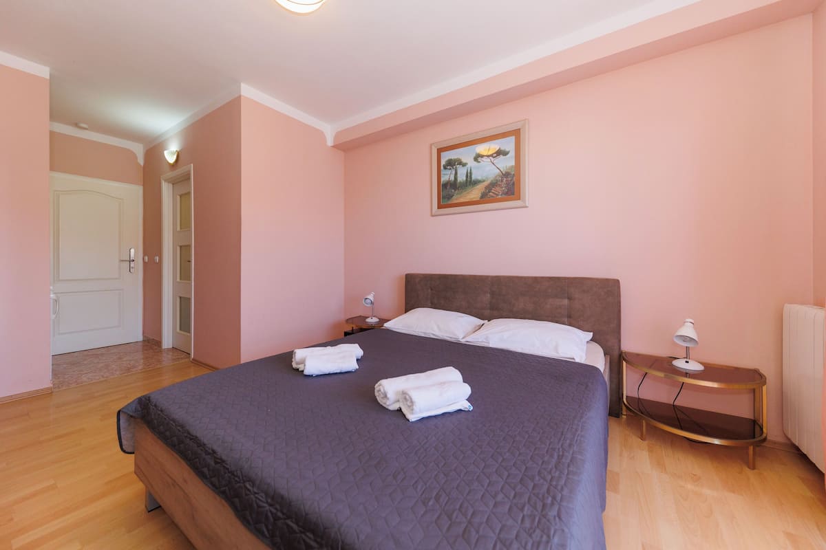 A bedroom with a large bed, two bedside tables, a nightstand, a painting on the wall, and a door leading to another room.