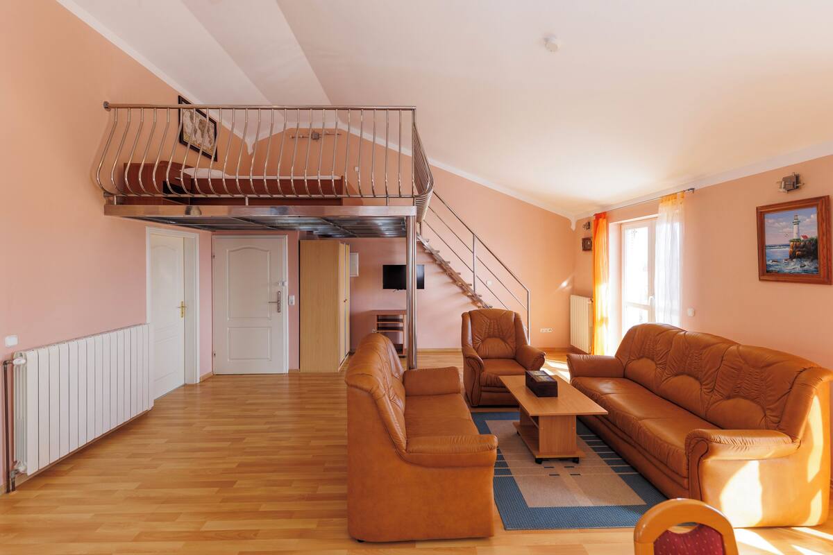 A spacious living area with a high ceiling, a wooden floor, a brown leather sofa, a wooden coffee table, and a staircase leading to an upper level.