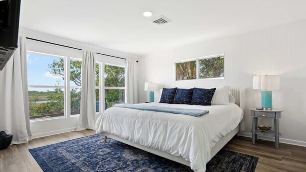 A bedroom with a large bed, a view of greenery outside, and a small bedside table with a lamp. - Waterfront Serenity, Steps to Beach, Fire Pit, BBQ (St. Augustine)