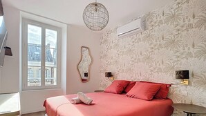 A bedroom with a large bed, a window with a view of buildings, a decorative mirror, and an air conditioning unit. - Amazon Suite (Montargis)