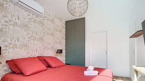 A bedroom with a bed, red bedding, a wall-mounted air conditioner, a patterned wallpaper, a door, and a hanging light fixture. - Amazon Suite (Montargis)