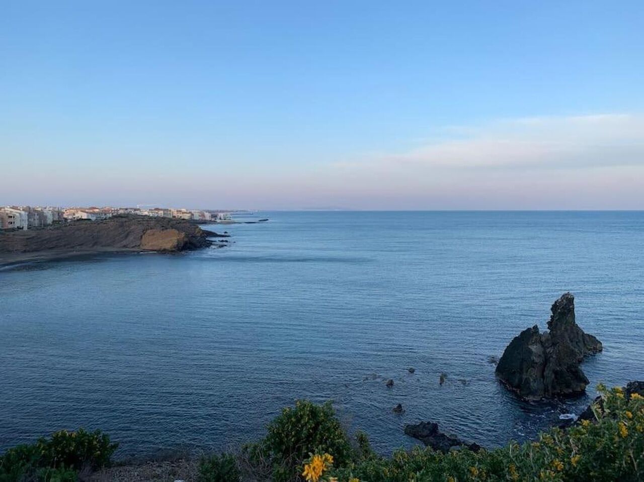A coastal landscape with a rocky outcrop in the water, a clear sky, and buildings along the shore.