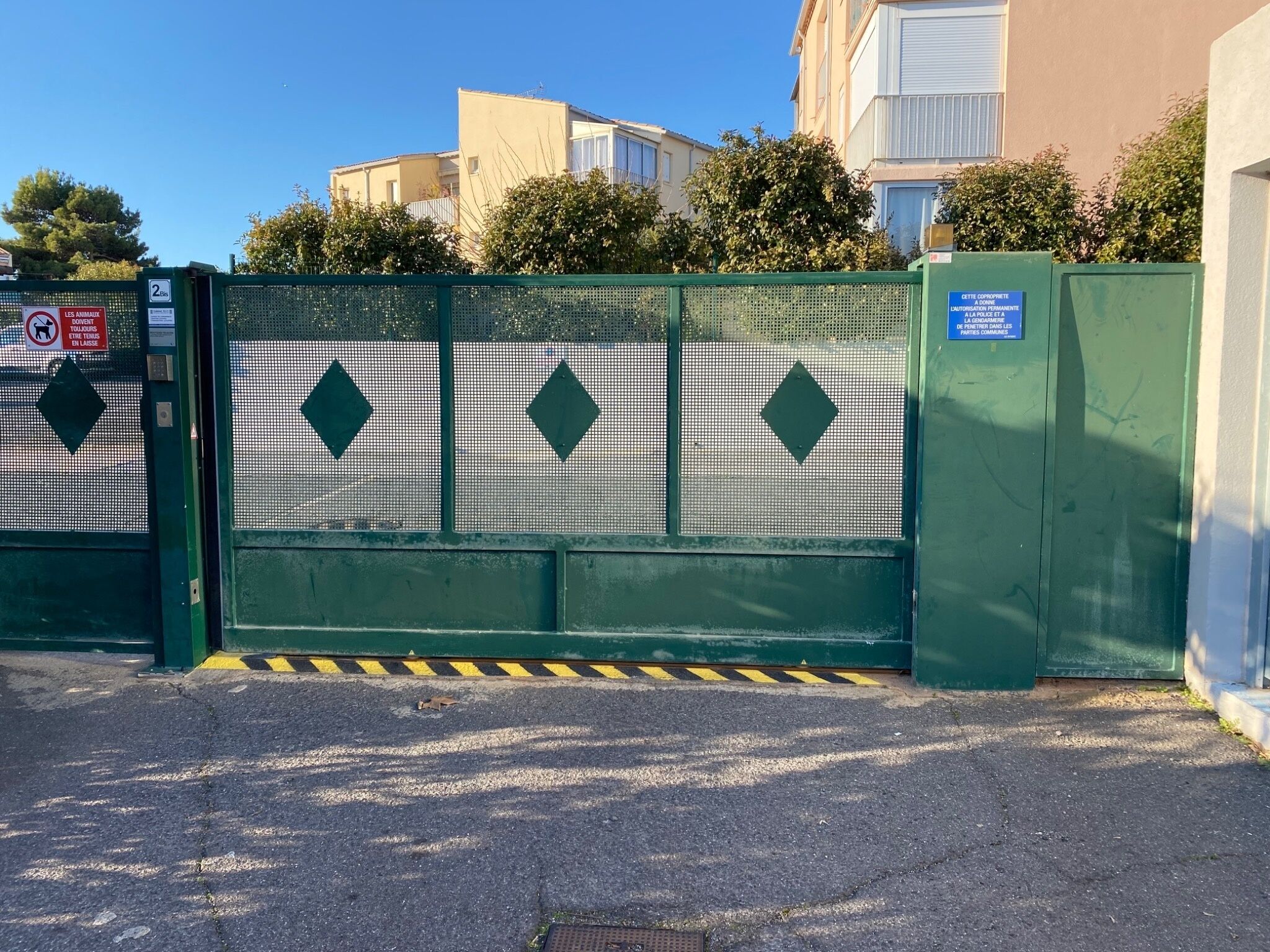 Une entrée fermée par un portail et une clôture en métal ornée de motifs en forme de losange, un panneau indiquant qu’il est interdit d’y accéder aux animaux, et un autre panneau signalant que le secteur est surveillé par la police.