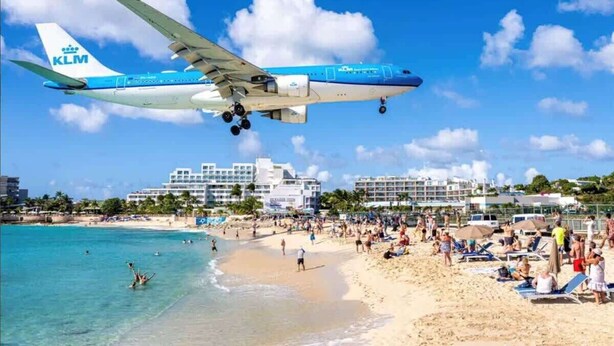 Una playa con un avión de KLM volando por encima, arena dorada y edificios al fondo.