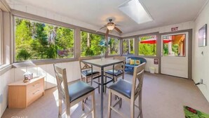 A sunlit room with a dining table, chairs, a ceiling fan, and large windows offering a view of greenery. An exit sign is visible on the wall. - The Lady Bug Family Retreat (Mariposa)