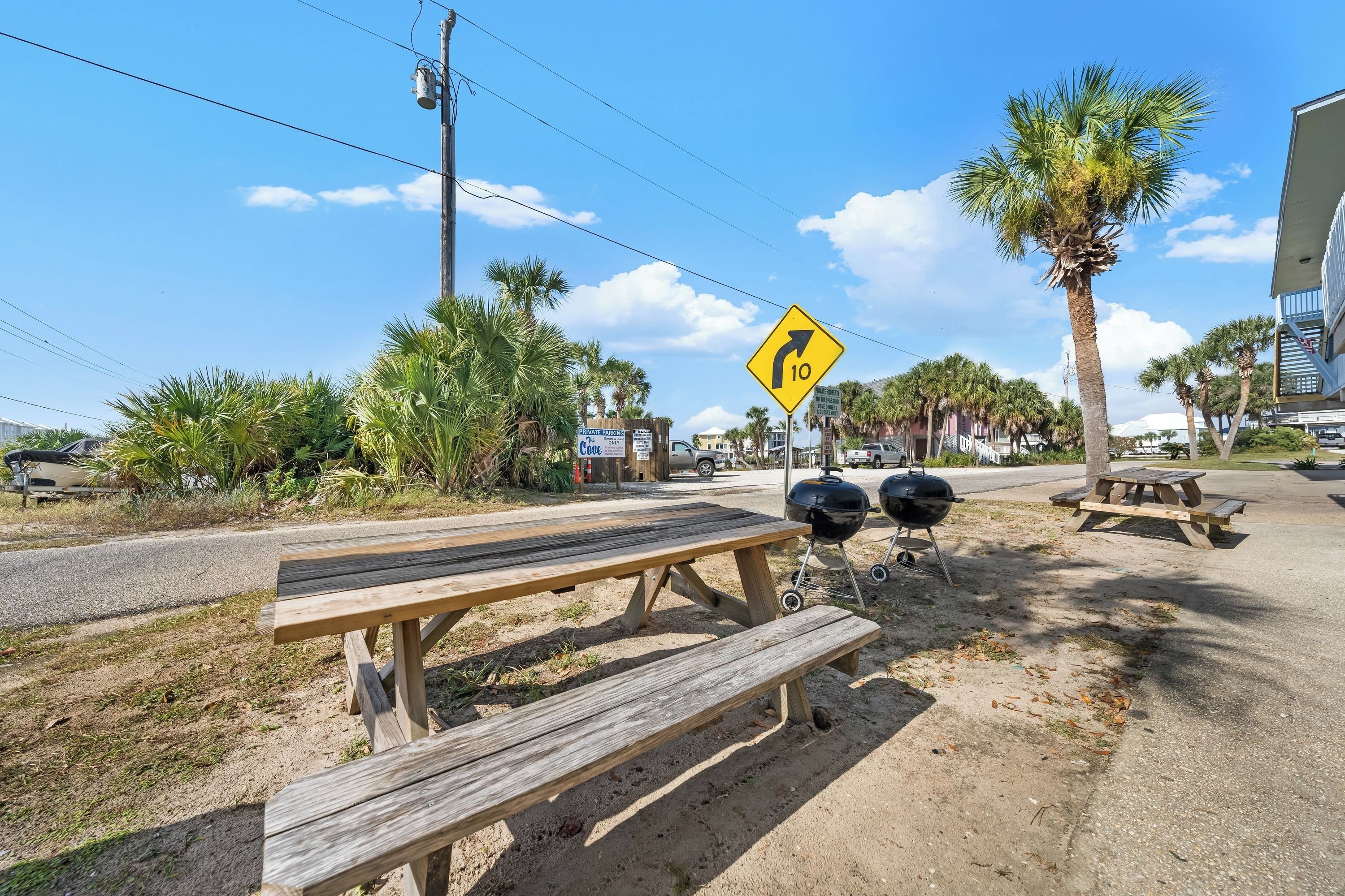 A roadside picnic area with a wooden bench and picnic table, a barbecue grill, and a road sign indicating a sharp turn.