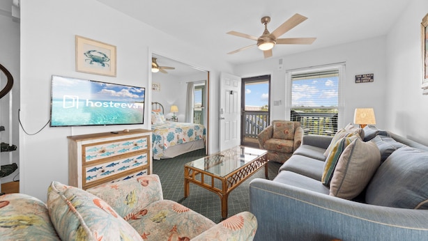 A living room with a television, a ceiling fan, a sofa, a chair, and a bed with a view.