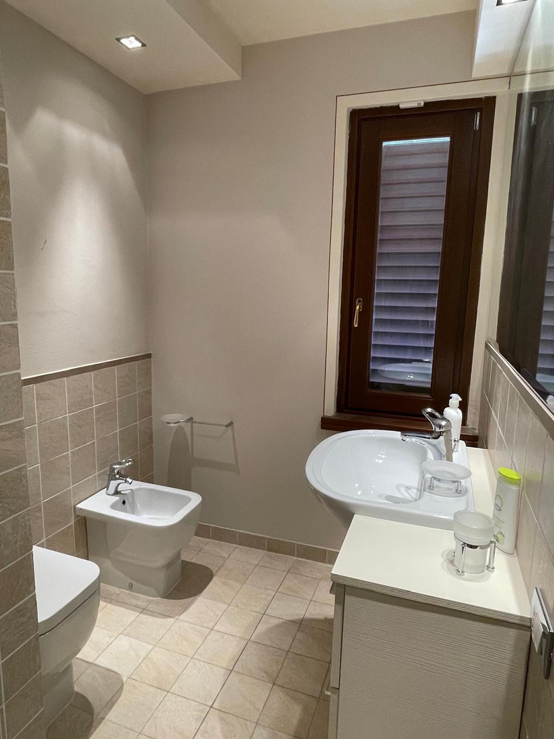 A bathroom with a bidet, toilet, sink, and a window with blinds.
