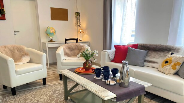 A modern living room with a white sofa, two armchairs, a coffee table, and a dining table with chairs. There is a globe on the dining table and a hanging plant above it.