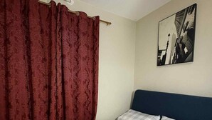 A bedroom with a bed, a checkered bedspread, a dark headboard, a red curtain, and a cityscape painting on the wall. - Fabulous building with AC in vibrant Cebu City (Cebu City)