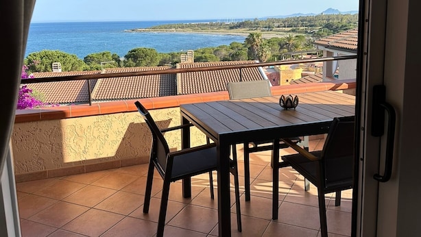 A balcony with a table and chairs, overlooking a coastal landscape with a clear view of the sea and distant mountains.