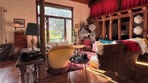 A cozy living room with a wooden ceiling, a large window, a sofa, a wicker chair, and a red curtain.