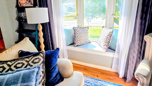 Un salon avec un canapé, des coussins à motifs, une lampe et une vue sur l’extérieur à travers une fenêtre avec des rideaux.