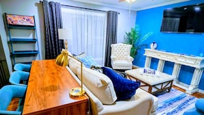 Un salon moderne avec une table basse en bois, un canapé blanc, des fauteuils bleus, un ventilateur de plafond et un téléviseur fixé au mur.