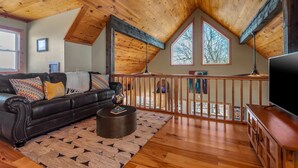 A cozy living room with a black leather sofa, a round coffee table, and a wooden TV stand. The room features a wooden ceiling with exposed beams, a balcony with a wooden railing, and large windows allowing natural light. - Winston's View: Mountain Views, Fire Pit, Hot Tub (Jasper)