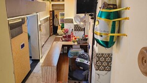 Un estrecho pasillo con una zona de cocina a un lado, un refrigerador, un ventilador y una pequeña mesa con flores.