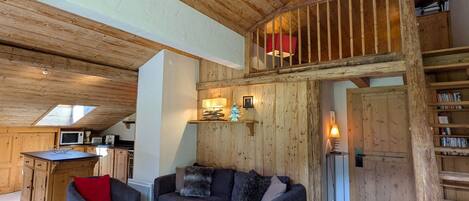 A cozy living area with a wooden ceiling, a sofa set, a coffee table, and a kitchen area with a microwave.
