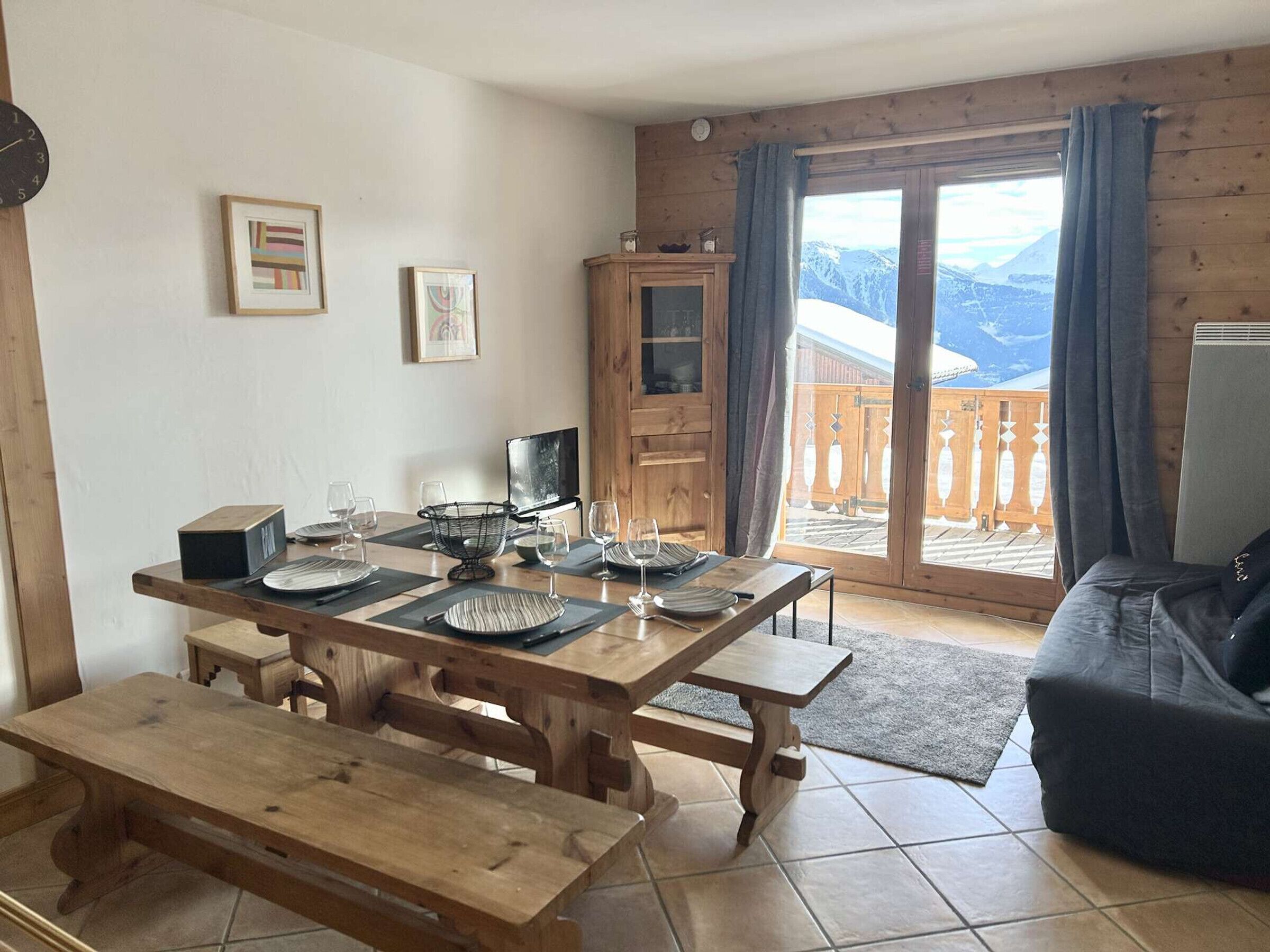 A wooden dining table set with plates, glasses, and cutlery, a wooden cabinet, and a view of mountains through a glass door.