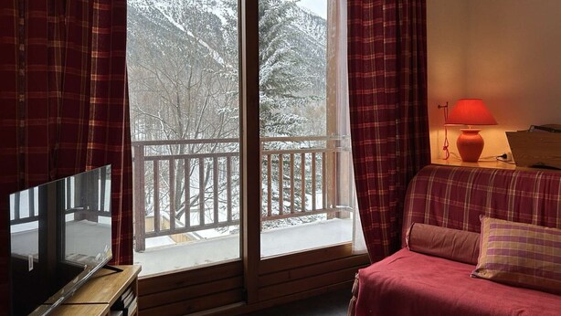 A room with a TV on a wooden stand, a sofa with a red blanket, a red lamp on a bedside table, and a view of snow-covered trees through a window with a wooden balcony. - Duplex chalet 6-8 ppl, garage, WiFi, Montgenèvre (Montgenèvre)