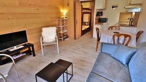 A cozy living room with a TV, sofa, and a small table. There's a wooden wall and a kitchen area with a microwave.