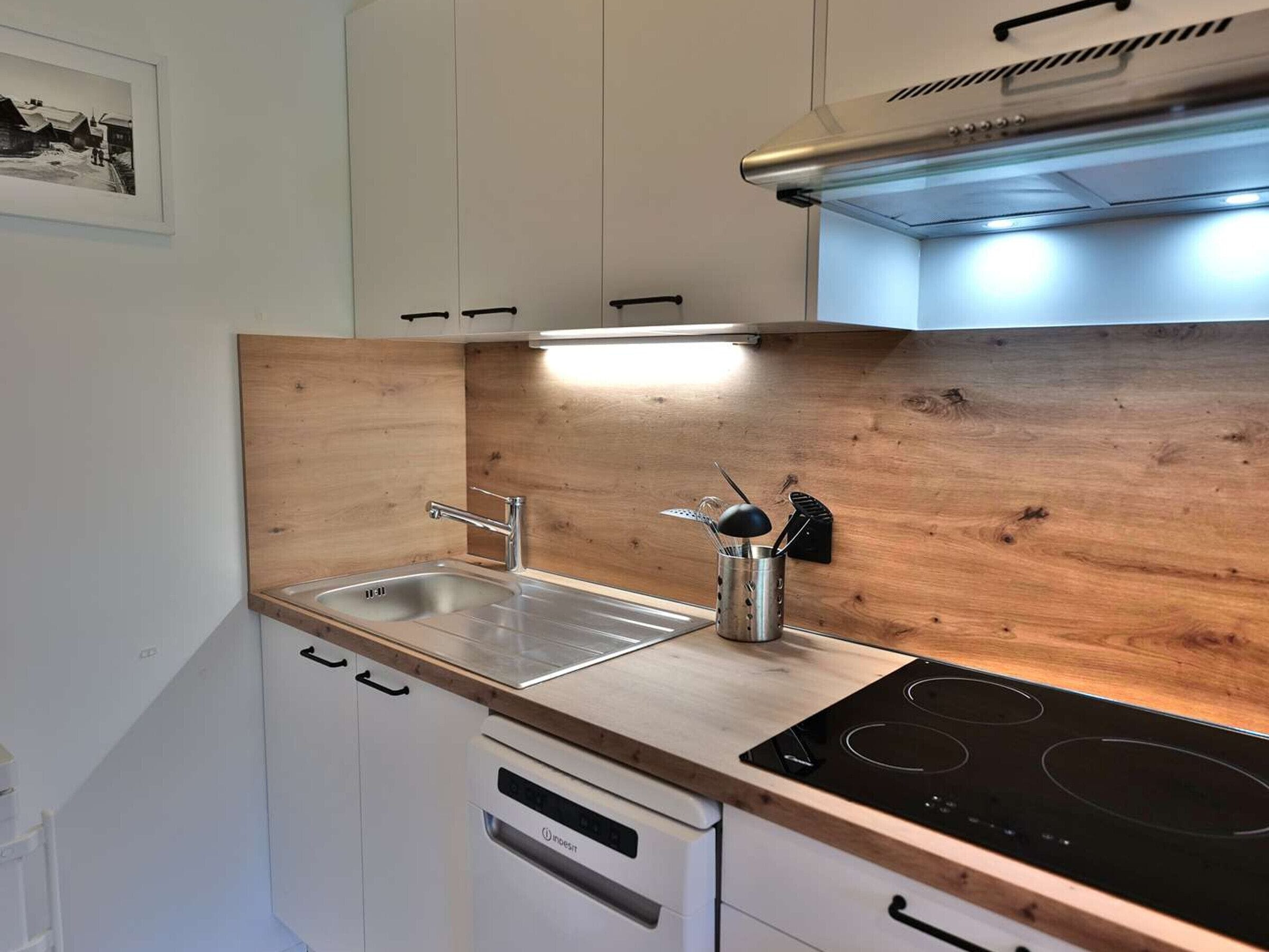 A compact kitchen with white cabinets, a stainless steel sink, a wooden backsplash, and a black induction cooktop.