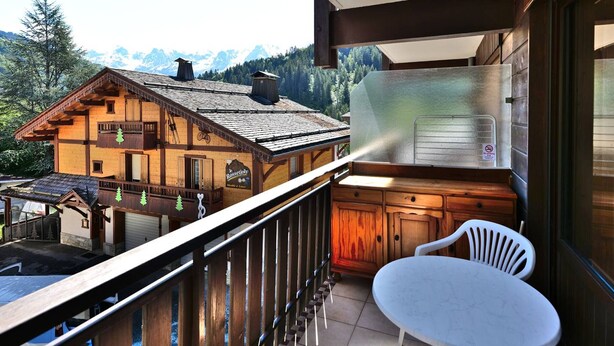 A wooden chalet with a balcony, a table, and chairs, overlooking a mountainous landscape.