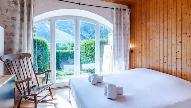 A bedroom with a wooden ceiling, a large bed with white linens, a rocking chair, and a view of greenery outside.
