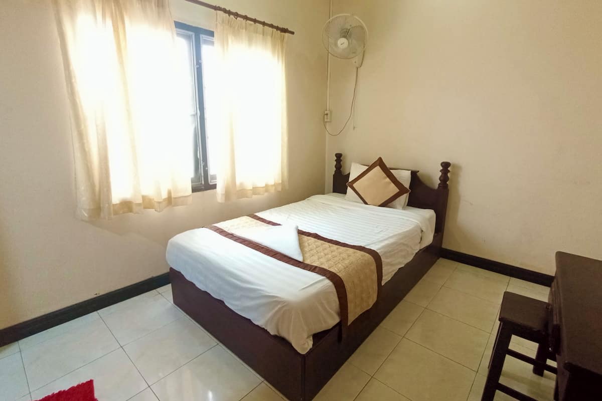 A bedroom with a bed, a window with curtains, a standing fan, and a small table.