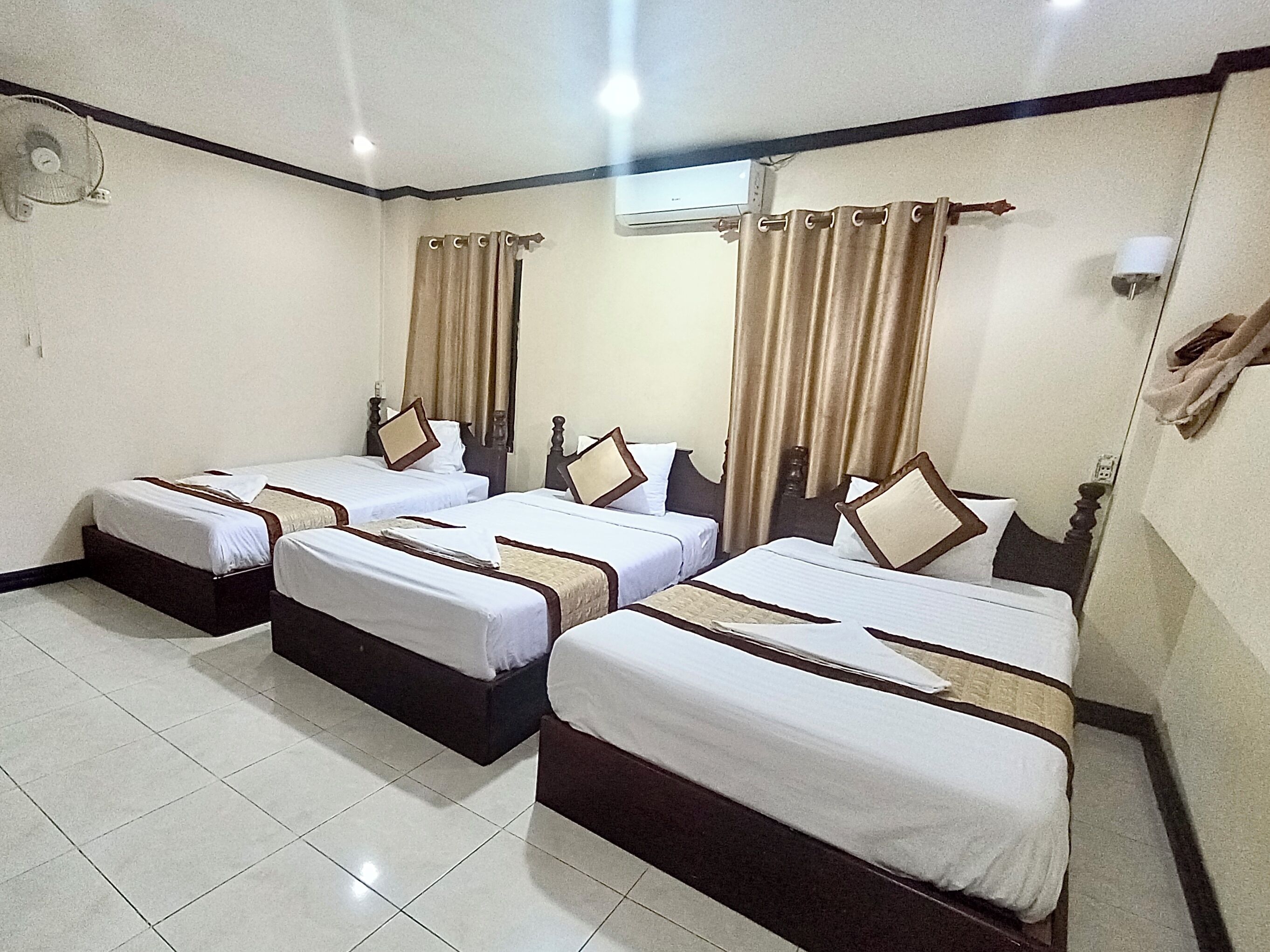 Habitación de hotel con tres camas individuales, cada una con cabecera de madera y ropa de cama blanca. El piso es de baldosas, las cortinas son color beige y tiene un ventilador de techo.