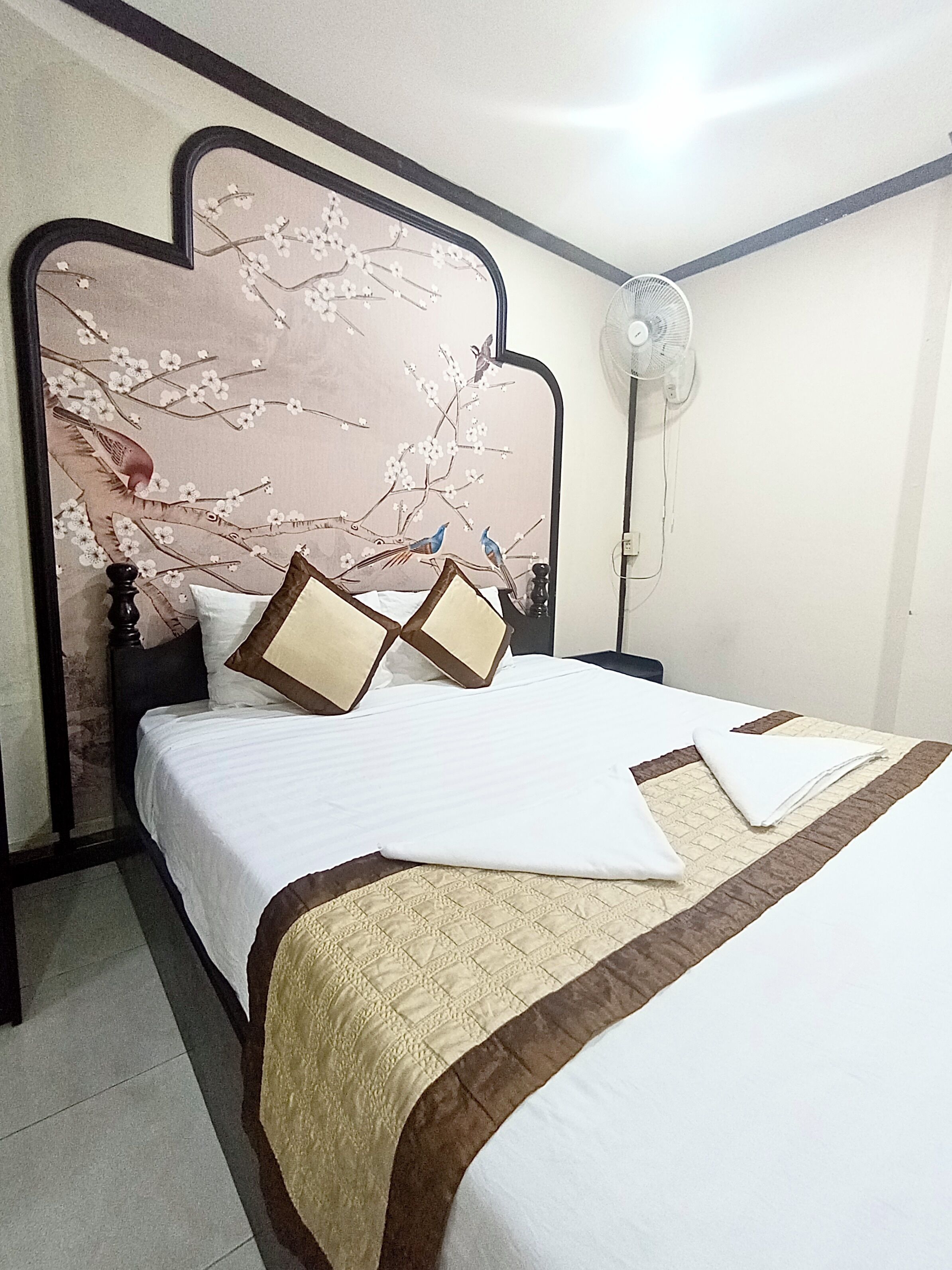 A neatly made bed with a white and brown bedspread, a headboard with a floral design, and a standing fan on the right.