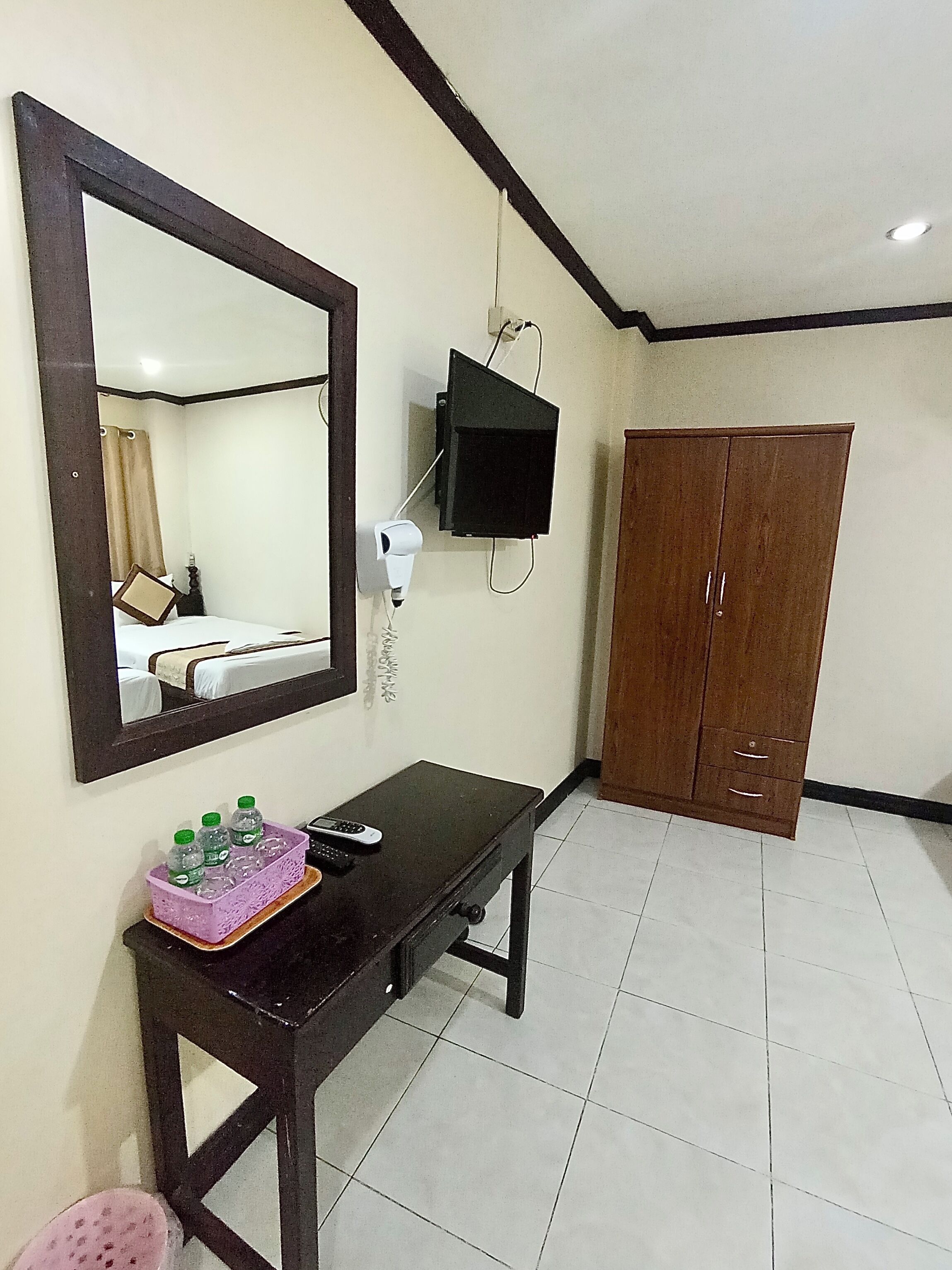 A hotel room with a large mirror, a TV mounted on the wall, a wooden wardrobe, a small table with toiletries, and a tiled floor.