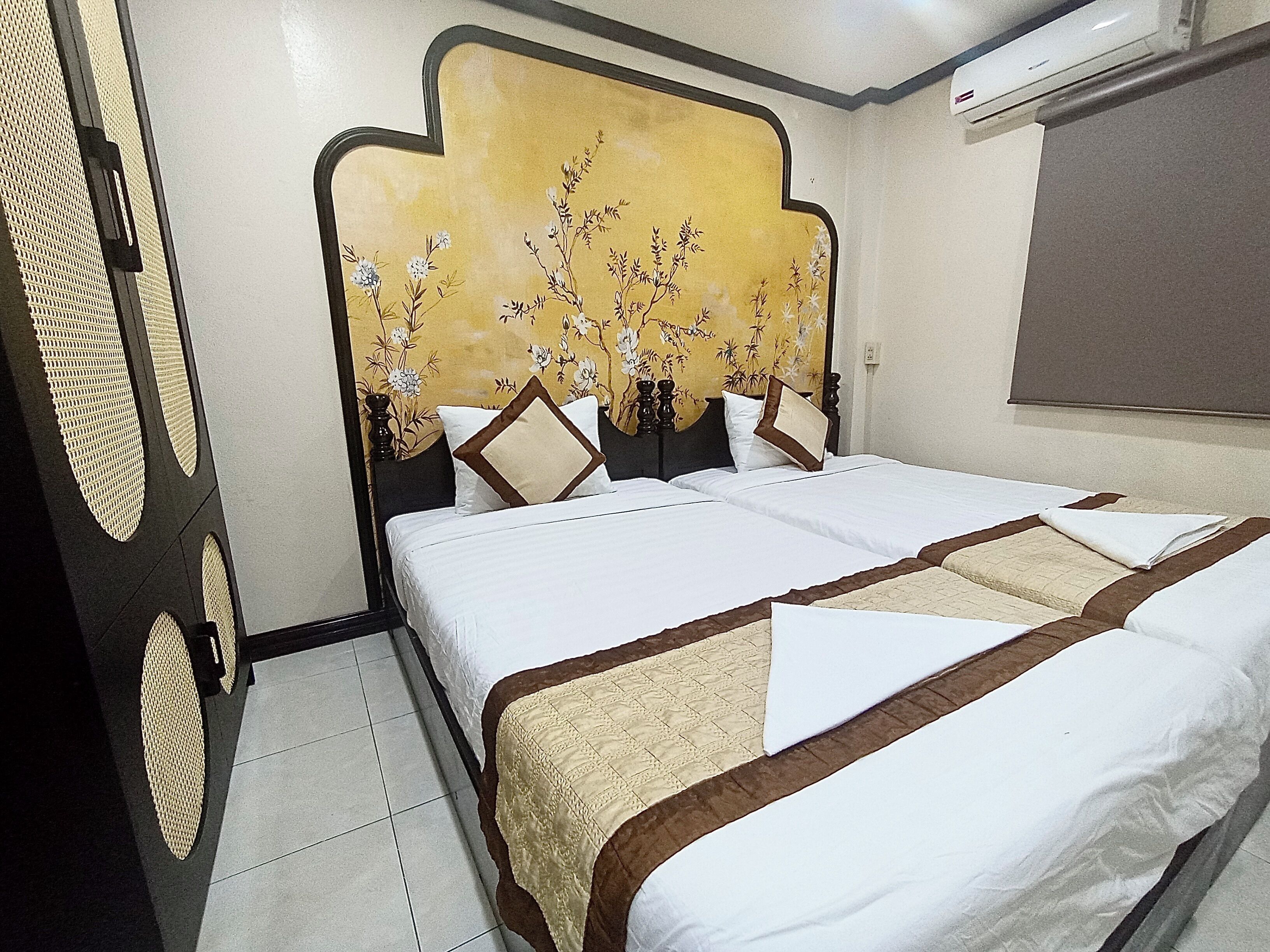 Une chambre d’hôtel avec un lit, une tête de lit ornée de motifs floraux et une fenêtre avec des stores.