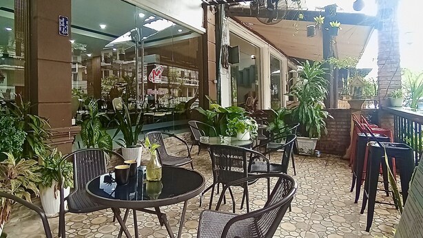 An outdoor seating area with round tables and wicker chairs, surrounded by potted plants and a tiled floor. - Memory Hotel Vientiane  (Vientiane)