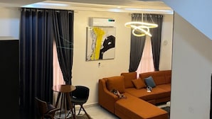 A modern living room with a sectional sofa, a small round table, and a chair. There is a painting on the wall and a ceiling light fixture. - R-Oak Hotels and Apartments  (Abuja)