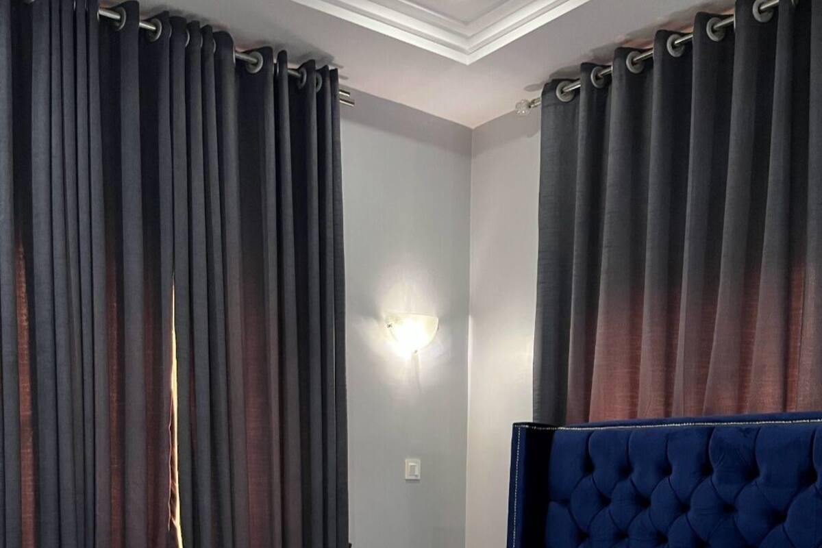 A hotel room with a large bed, dark curtains, a wall-mounted light, and a blue headboard.