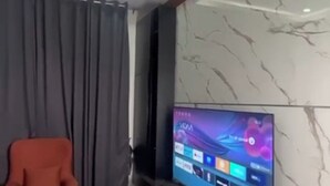 A modern living room with a flat-screen TV on a wooden stand, grey curtains, a red chair, and a marble wall. - R-Oak Hotels and Apartments  (Abuja)