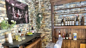 Angolo bar con parete in mattoni, ripiano in legno con bottiglie, una poltrona imbottita e un bancone con un vaso di fiori.