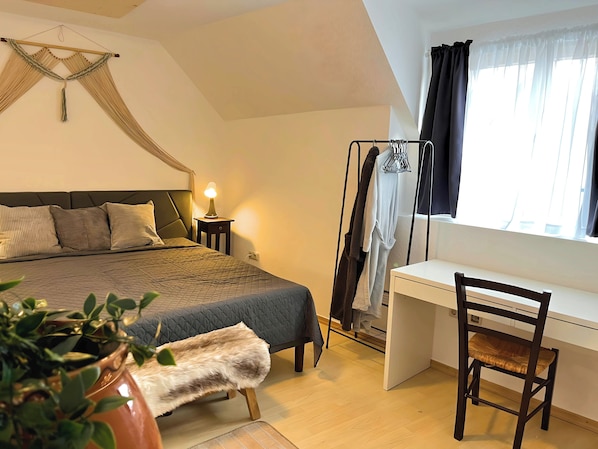 A bedroom with a bed, bedside table, lamp, hanging clothes rack, desk, chair, and a potted plant.