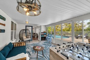 A spacious living area with a foosball table, a hanging chair, and a view of a backyard with a pool. - Modern 4BR Home w/ Private Heated Pool & Firepit (Seminole)